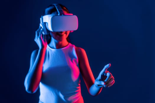 Smart female stand with surrounded by cyberpunk neon light wear VR headset connecting metaverse, futuristic cyberspace community technology. Woman using finger pointing virtual object. Hallucination.
