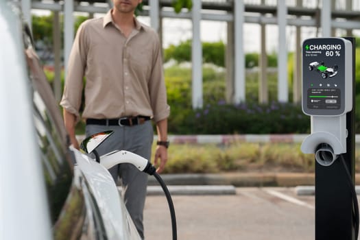 Young man put EV charger to recharge electric car's battery from charging station in city commercial parking lot. Rechargeable EV car for sustainable environmental friendly urban travel. Expedient