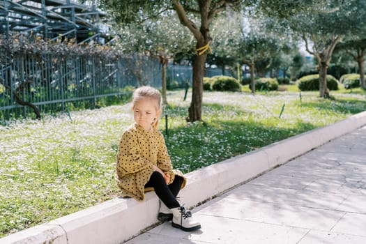 Little girl sits on the curb near a flowering meadow and looks away. High quality photo