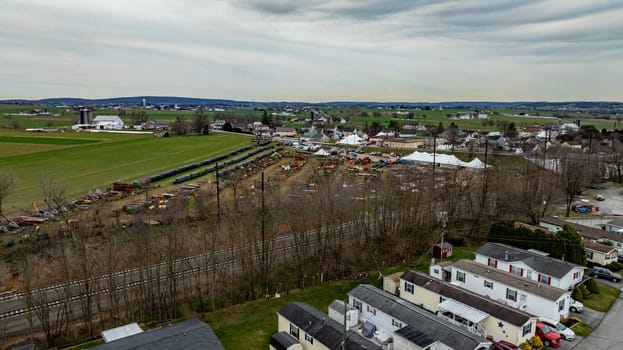 A drone photo capturing a lively rural community with residential areas, agricultural machinery, and a small fair setup under an overcast sky.