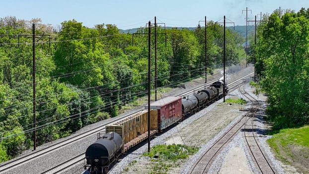 An engaging aerial view of a freight train carrying diverse cargo winding through rural tracks surrounded by lush greenery, under a bright sunny sky.