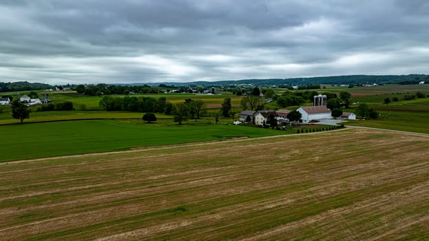 This image captures the sprawling beauty of a rural farmstead surrounded by a patchwork of lush green and harvested fields. The overcast sky casts a serene glow over the farm buildings