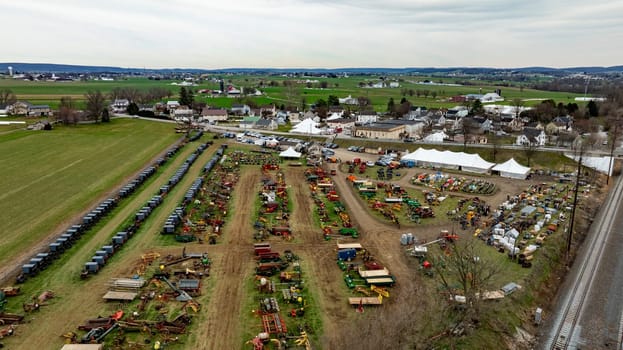 This Aerial image of an Amish Mud Sale, shows a comprehensive display of colorful farm machinery and equipment spread across a rural exhibition field with tents and visitors.