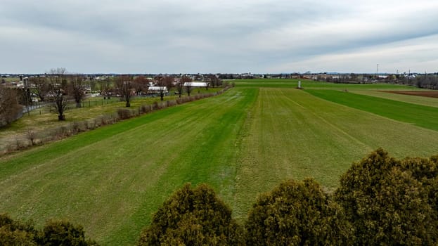 This drone-captured image showcases a vast rural landscape with distinct green fields, a few clusters of trees, and a small town in the background under a cloudy sky.