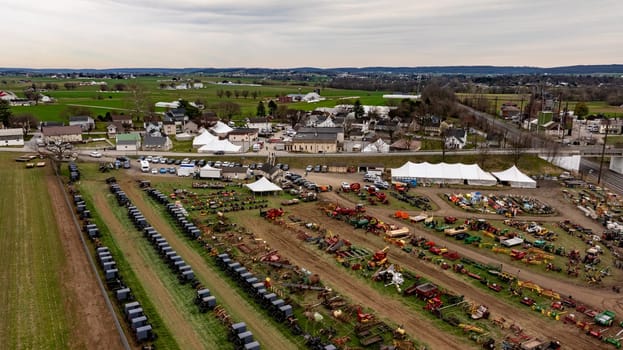 This Aerial image of an Amish Mud Sale, shows a comprehensive display of colorful farm machinery and equipment spread across a rural exhibition field with tents and visitors.