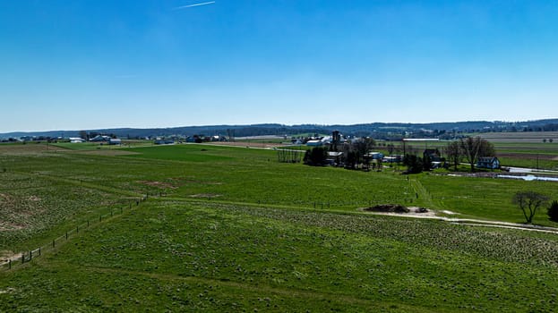 An expansive view from above showcasing lush green pastures segmented by fencing, with a detailed farmstead and distant hills under a brilliant blue sky.