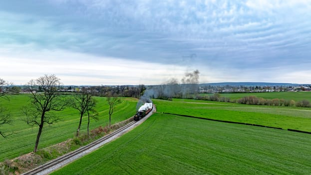 Dramatic aerial capture of a vintage steam train chugging along a curvy track through lush green fields, with a backdrop of dramatic cloudy skies and a distant village.