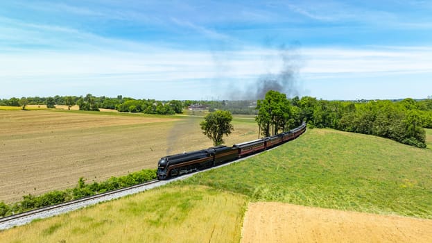 Captivating aerial view of a vintage steam train traveling through a vast rural landscape, with plowed and green fields under a blue sky.