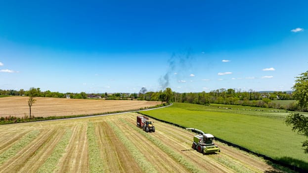 An aerial shot displaying intense harvesting activity with machinery in action across a multi-textured landscape of cut and uncut fields, under a bright blue sky with distant smoke visible.