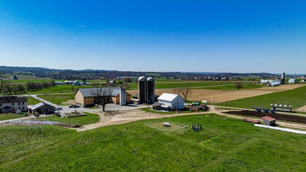 Overhead shot of a busy farm complex, featuring multiple barns and silos surrounded by verdant pastures, farm equipment in use, and a group of people gathered, under a clear sky.