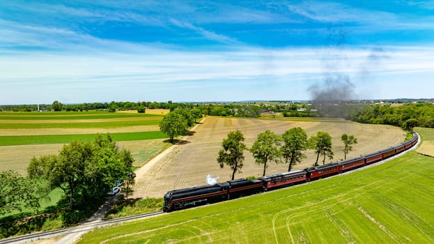 Aerial image of a classic steam train curving through the countryside, emitting smoke above vibrant and plowed fields under a blue sky.