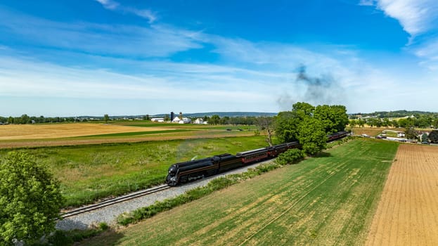 A striking aerial view showcasing a historic train cutting across scenic farmlands, with mixed agriculture fields and a backdrop of blue sky.