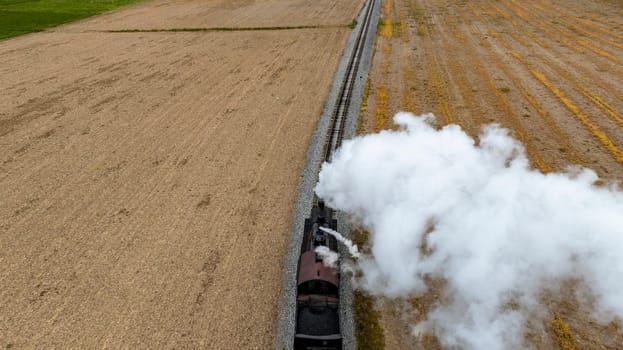 Dramatic top-down view of a steam train emitting a thick cloud of smoke as it travels alongside freshly harvested fields, highlighting the contrast between technology and nature.