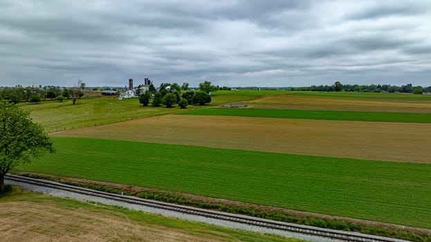 This aerial photograph highlights a serene farmland, featuring vibrant green patches of crops, separated by the diagonal sweep of a railroad track. The farm's infrastructure, including silos and barns