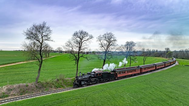 Eye-catching aerial image of a classic steam locomotive pulling red passenger cars through a lush green countryside, with trees and a cloudy sky adding to the scenic beauty.
