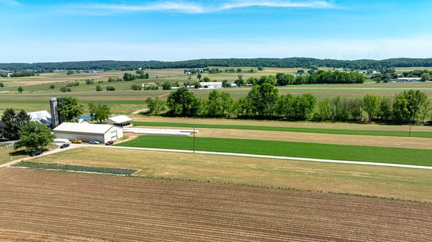 An expansive aerial shot capturing the serene beauty of a rural farm, showcasing a mix of plowed fields, green crops, and farm buildings under a clear blue sky.