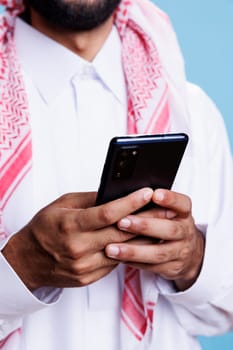 Arab man holding mobile phone in hands, scrolling social media network and texting online closeup. Muslim person in traditional clothes using smartphone and surfing internet