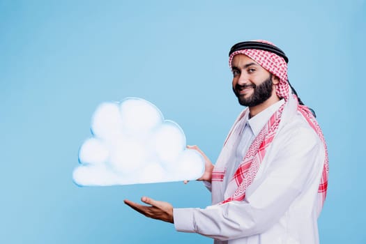 Cheerful man dressed in muslim traditional clothes showing white cloud shape and looking at camera with smiling expression. Carefree arab holding cardboard in studio portrait on blue background