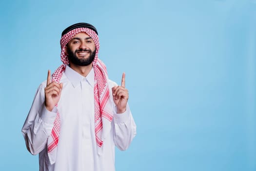 Excited muslim sales person pointing upwards direction while posing for studio portrait. Cheerful smiling arab man wearing traditional clothes showing up and looking at camera