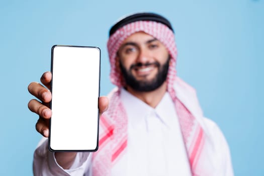 Man in arabic attire holding smartphone displaying white screen for application advertising. Smiling muslim person posing in studio and showing mobile phone blank touchscreen closeup