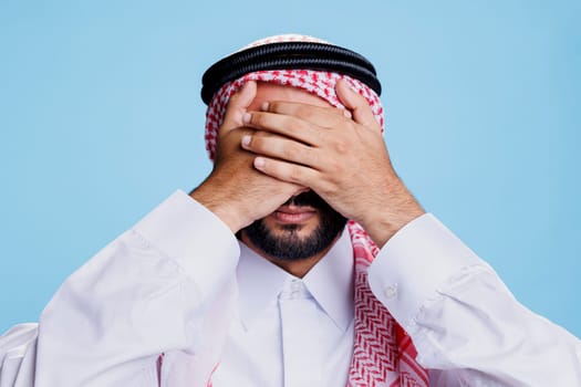 Muslim man covering eyes with palms while showing see no evil three wise monkeys concept. Person wearing traditional arabic clothes showcasing hide and seek gesture in studio