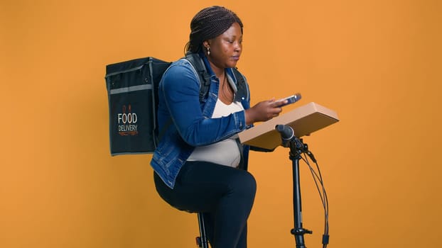 Black woman delivers takeout orders on bicycle, utilizing wireless payment device for seamless transactions. African american delivery person taking pos machine from pocket for contactless pay.