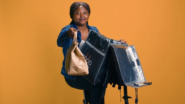 Reliable courier showing thumbs up after delivering takeout meal on bicycle as eco-friendly transportation. Active african american lady providing exceptional delivery service of hot fast food.