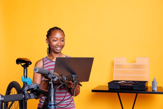 African American female cyclist repairs broken bike utilizing toolbox and personal computer to browse for solutions. Young black woman working on modern bicycle by using minicomputer and expert tools.