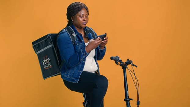 Professional african american cyclist uses bicycle and smartphone to offer efficient food delivery service. Young black woman embraces fast-paced economy of utlizing mobile device for courier job.