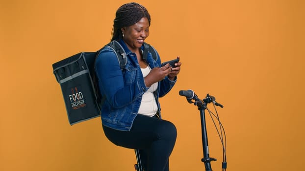 Female courier relaxing with smartphone on bicycle on break from delivery service. Enthusiastic african american woman checking cell phone while waiting for online restuarant order for delivering.