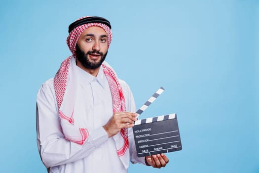 Muslim man dressed in thobe and headscarf clapping movie slate, showing film scene action studio portrait. Arab person wearing traditional clothes holding clapperboard and looking at camera