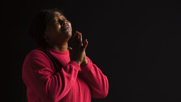 Graceful black woman gazing upwards with palms extended in gesture of reverance and adoration. Side-view portrait of african american lady engaged in prayerful reflection in isolated background.