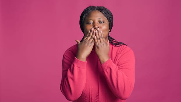 Lady with african american ethnicity blowing a kiss towards camera being lovely isolated on pink background. Stunning joyful black woman expressing love with cute kissing hand gestures.