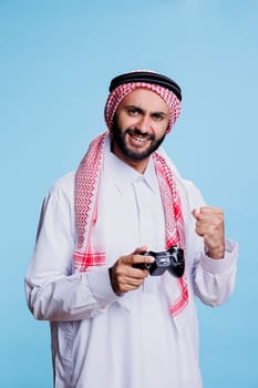 Excited muslim man holding joystick and showing clenched fist while winning game studio portrait. Happy person dressed in arabic traditional clothes posing with gamepad and winner gesture