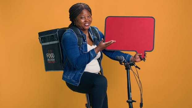Enthusiastic black woman with food delivery bag on back, sitting and gesturing on red sign in her hand. Friendly courier holding red speech bubble, showcasing reliable eco-friendly delivery app.