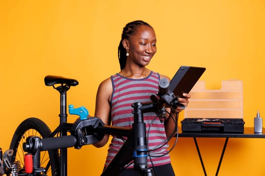 Sports-loving black woman inspecting broken bicycle, examining damaged components, researching repair solutions on tablet. African american female grasping device for bike repair online instructions.