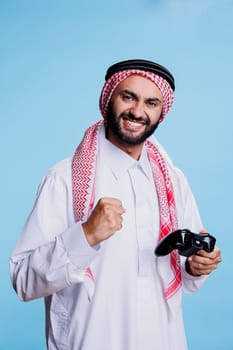 Happy muslim man showing winner gesture with clenched fist and holding wireless joystick studio portrait. Arab person wearing traditional standing with console gamepad and winning videogame