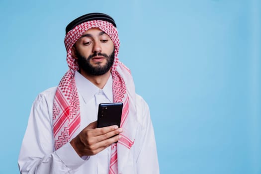Muslim man wearing traditional clothes texting on smartphone while posing in studio. Arab person dressed in thobe and ghutra sending online message on mobile phone on blue background