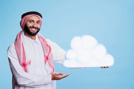 Carefree man wearing arabic traditional clothes posing with cloud shape cardboard banner studio portrait. Smiling muslim person holding showing white bubble and looking at camera