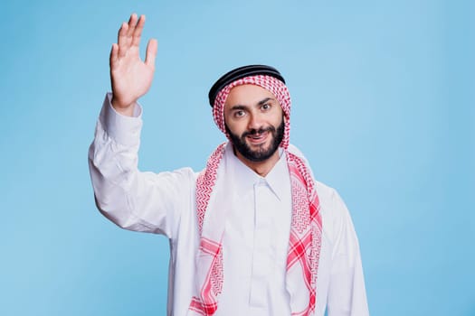 Smiling muslim man dressed in traditional clothes raising hand studio portrait. Islamic person greeting friend, waving with arm and looking at camera with cheerful expression
