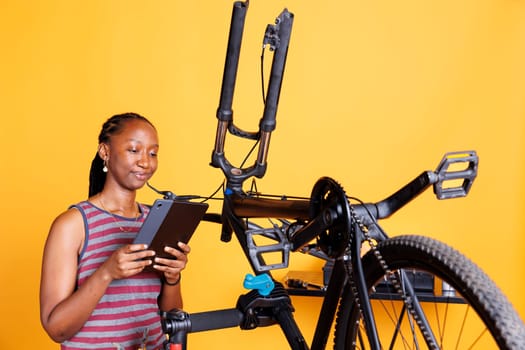 Image showing african american female holding a tablet and inspecting damaged bike components. Active sporty black woman using smart digital device to research bike repair instructions.