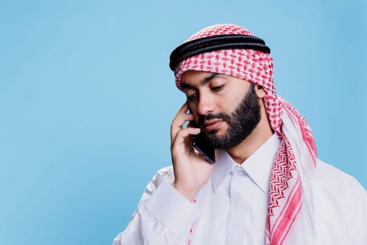 Man wearing arab thobe and headscarf speaking on mobile phone with neutral face expression. Muslim person in islamic headdress speaking on smartphone while posing in studio