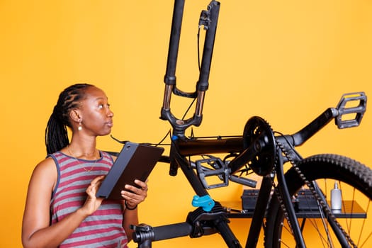 Healthy African American lady fixing bicycle with tools and browsing tablet for solutions. Youthful black woman carefully examines and repairs bike frame using repair-stand and digital device.