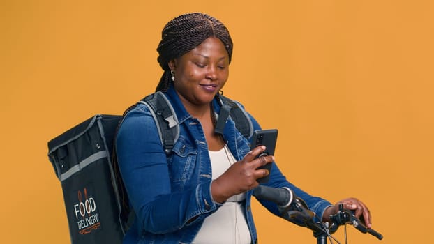 Active black woman delivering food on bicycle with cell phone in hand. African american female checking delivery app on smartphone for transportation of package. Portrait shot, smiling at camera.