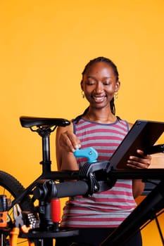 Active black woman examining damaged bicycle parts and using digital device for repair research. Sporty female multitasking, servicing bike and utilizing her digital tablet for guidance.