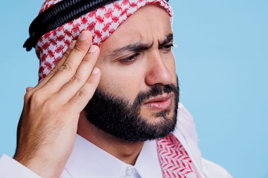 Muslim man rubbing temple and suffering from migraine symptom with tired expression. Arab person wearing thobe and ghutra traditional clothes having headache and feeling pain
