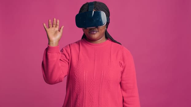 Innovative young black woman enjoys futuristic simulation with interactive device while wearing 3D virtual reality glasses in studio. African american trendy fashionista using modern VR headset.