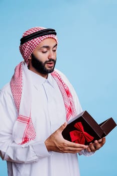 Muslim man wearing traditional attire holding giftbox with bow ribbon and opening festive present. Arab dressed in white thobe standing with giftbox in studio on blue background