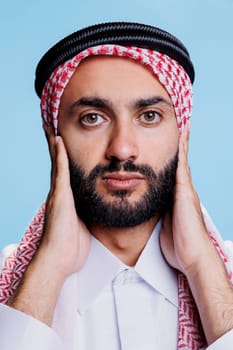 Man wearing muslim checkered headscarf covering ears with arms and looking at camera closeup. Arab person wearing traditional islamic clothes showing hear no evil three wise monkeys concept