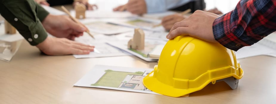 Professional architect team and engineer discuss about architectural project on meeting table with safety helmet, wooden block and architectural document scatter around. Closeup. Delineation.